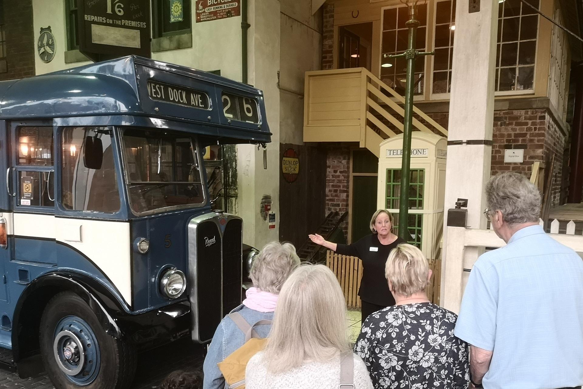 A woman stood next to a bus talking about it to a group of people facing her.