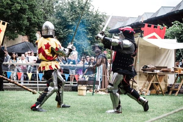 Two people in Knights costume fighting with swords. There are people and tents in the background