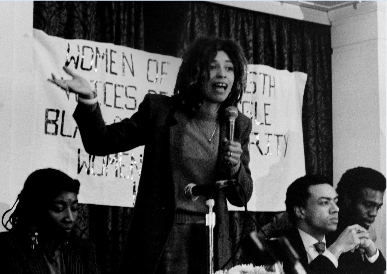 A black and white photograph of a woman speaking at an event.